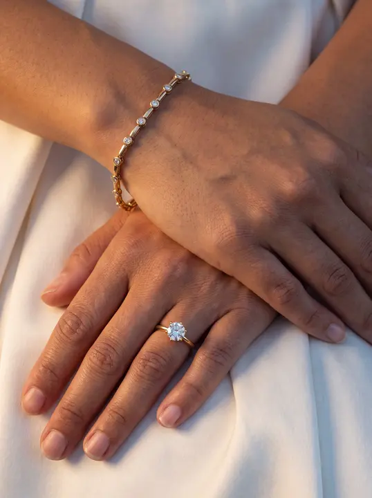 Hands crossed on white fabric, thin diamond bracelet and solitaire ring in gold.