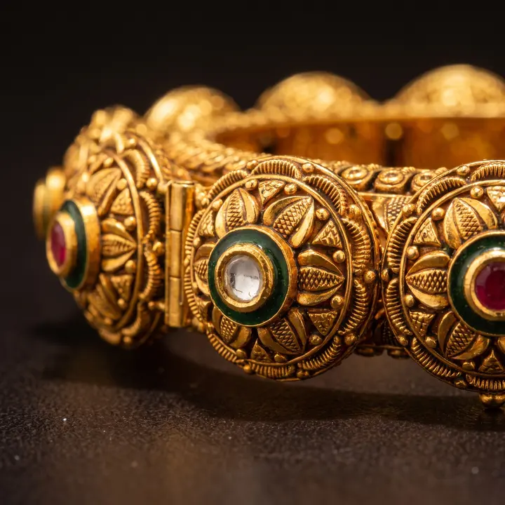 Plain gold kada with granulated detail and green enamel stones on a dark ground, low angle.