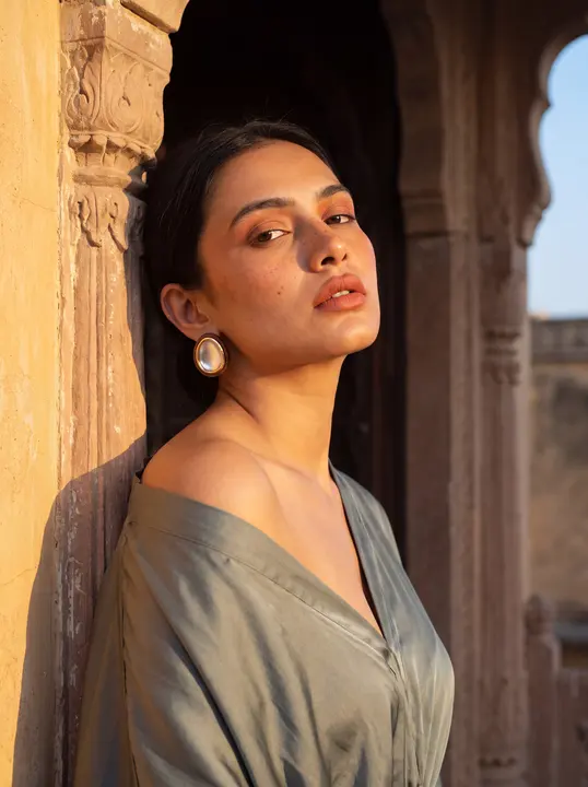 Woman leaning in a sandstone arched doorway, sage-green top, pearl drop earring; the identity reference portrait.