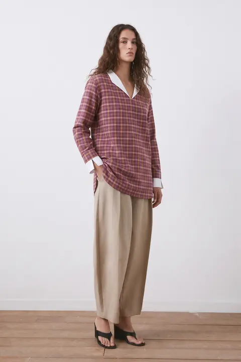 Woman in the Sabki Madras shirt worn loose over wide beige trousers and sandals, light grey studio wall and wood floor.
